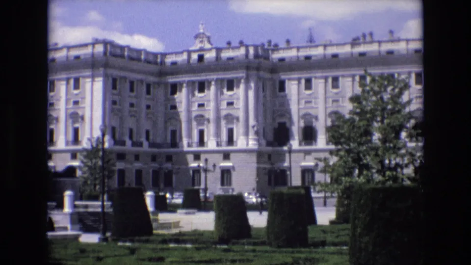 1972 Spain 8mm frame focused on a large pale palace facade across a manicured garden, with clipped hedges, trees, lampposts, and small distant visitors visible near the walkway.