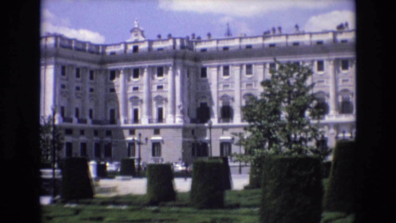 1972 Spain 8mm frame showing a grand palace exterior with rows of windows, a roofline balustrade, clipped hedges, trees, and a paved foreground.