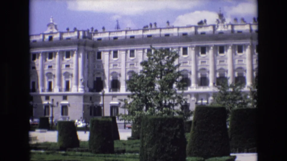 1972 Spain 8mm frame of a large classical palace seen from garden grounds, with trimmed hedges, trees, many windows, and a pale blue sky with clouds.