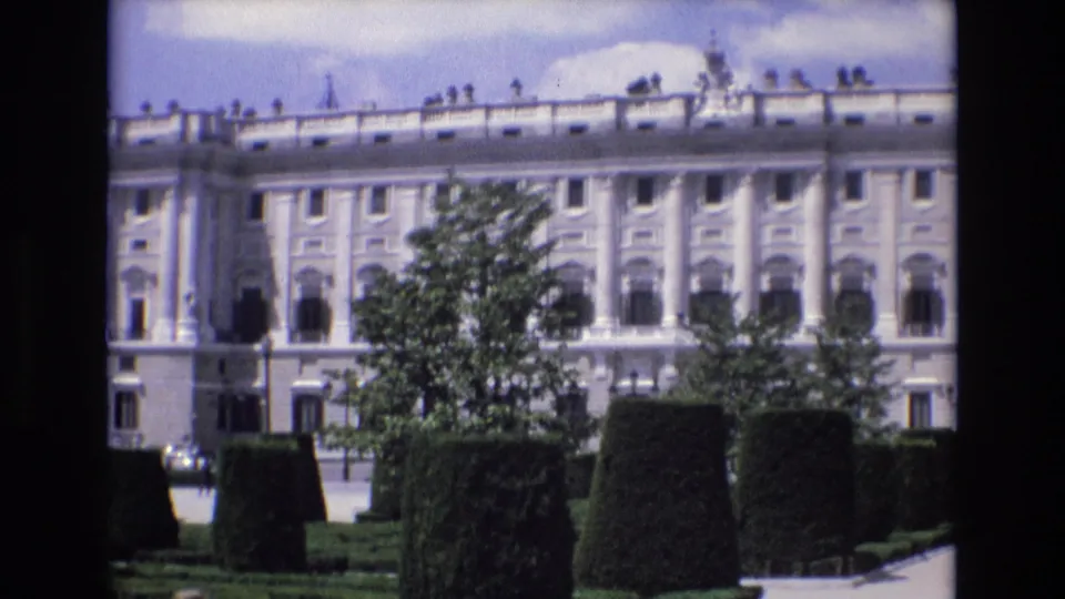 1972 Spain 8mm frame holding on the palace wall and windows, with a leafy tree and clipped garden hedges partially screening the lower facade.