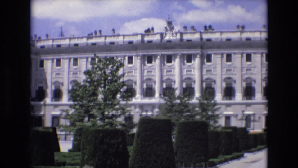 1972 Spain 8mm frame showing a wide exterior view of the palace and manicured grounds, with clipped shrubs, trees, repeated windows, and distant visitors near the building.