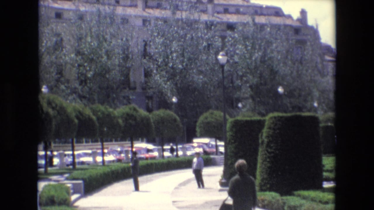 Pedestrians in a landscaped Spanish public garden walk along a curved path with trimmed hedges, street lamps, trees, and cars beyond the greenery.