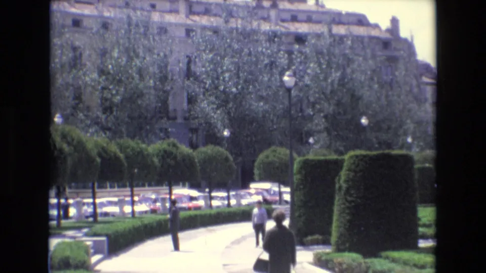 Two distant pedestrians walk through a formal garden walkway in Spain, with shaped hedges, leafy trees, street lamps, parked or passing cars, and a large building behind them.