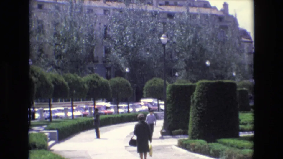 A woman carrying a bag and another pedestrian walk along a curved landscaped path in Spain, with trimmed hedges, mature trees, street lamps, cars, and a large building visible beyond.