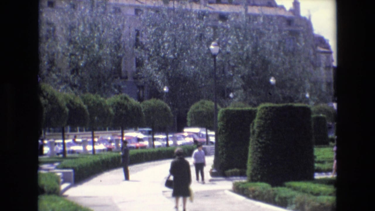 In Spain archival 8mm footage from 1972, pedestrians walk through a formal landscaped park with clipped hedges, trees, street lamps, and parked cars beyond the path.