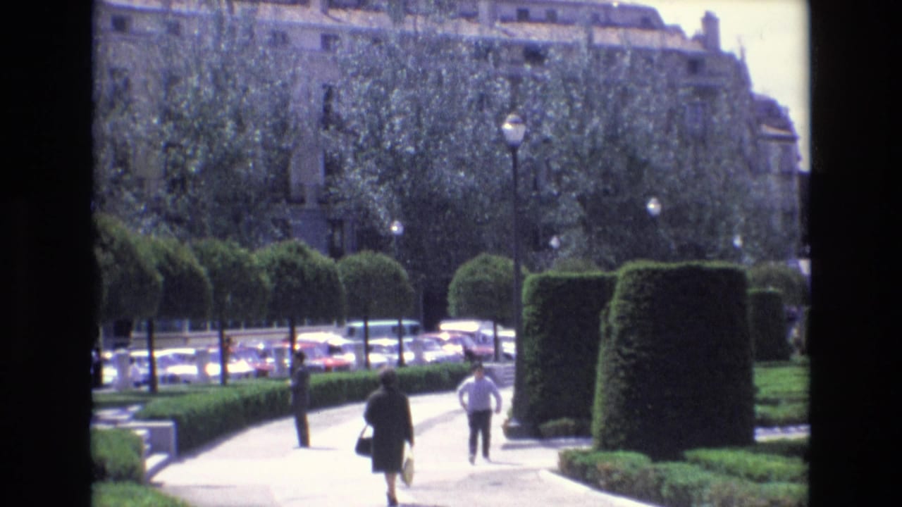 Two pedestrians walk on a sunlit path between sculpted hedges in a formal park.