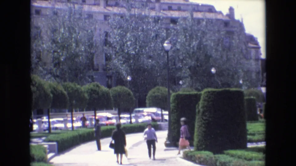 This Spain 1972 8mm still shows visitors walking in a landscaped public park, with clipped hedges, dense trees, globe street lamps, and parked cars visible beyond the walkway.