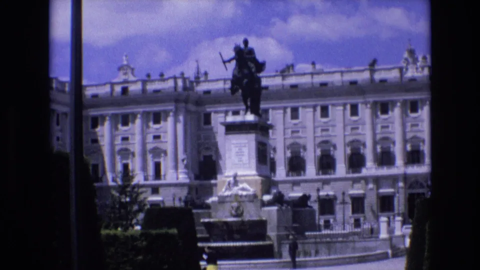1972 Spain home-movie frame showing a grand palace facade, an equestrian statue on a pedestal, clipped greenery, and a fountain-like stone feature in the foreground.