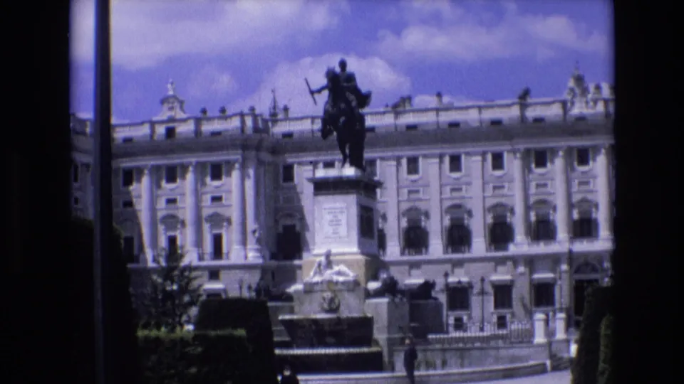 1972 Spain home-movie view of a palace plaza with an equestrian monument, pale stone facade, fountain-like feature, trimmed planting, and a distant pedestrian.