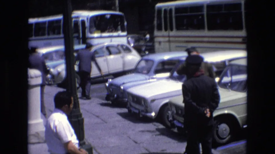 1972 Spain street-side frame showing parked vintage cars, buses, pedestrians, and a uniformed figure beside a lamppost near the palace area.
