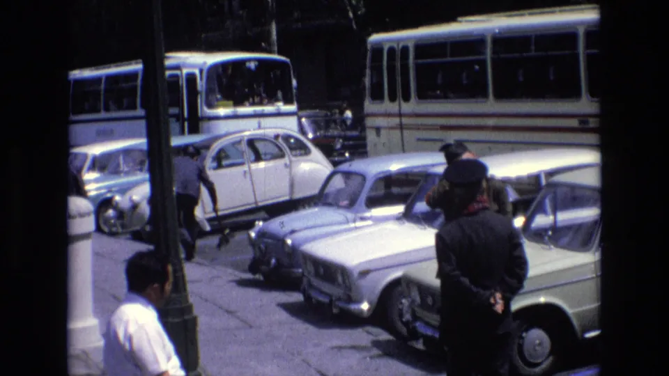 1972 Spain home-movie frame showing adult men near parked classic cars, with public buses and a lamp post visible in the background.