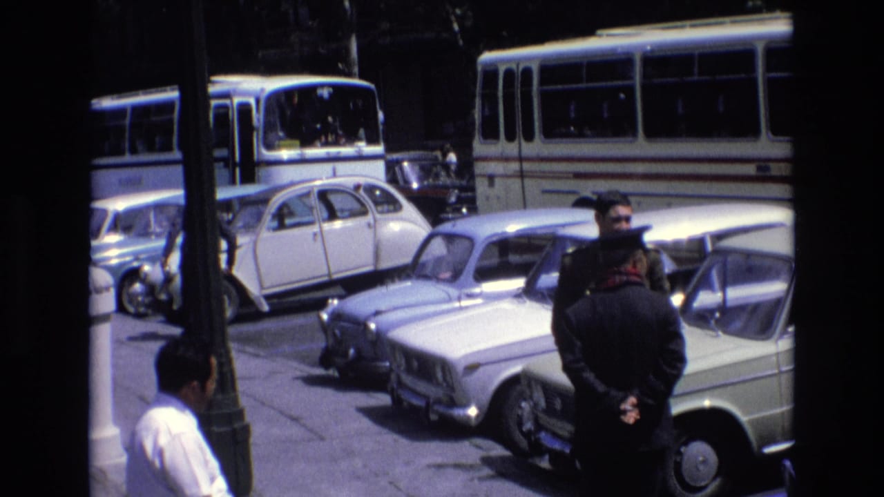 A uniformed-looking man faces another man near parked cars and buses.