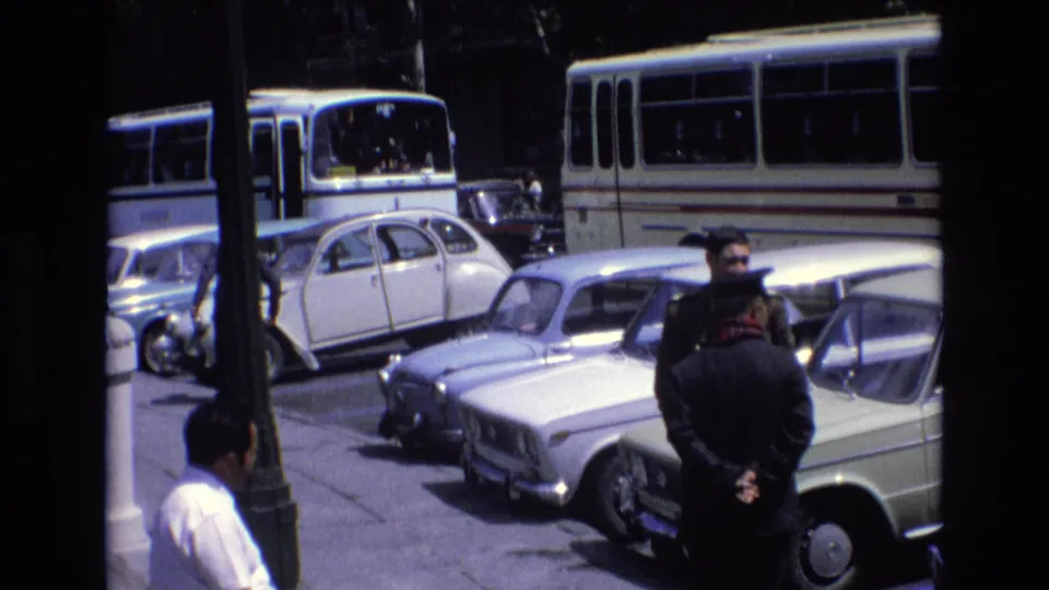 1972 Spain street frame with a uniformed-looking adult man and another man standing among parked cars, buses, and sidewalk fixtures.