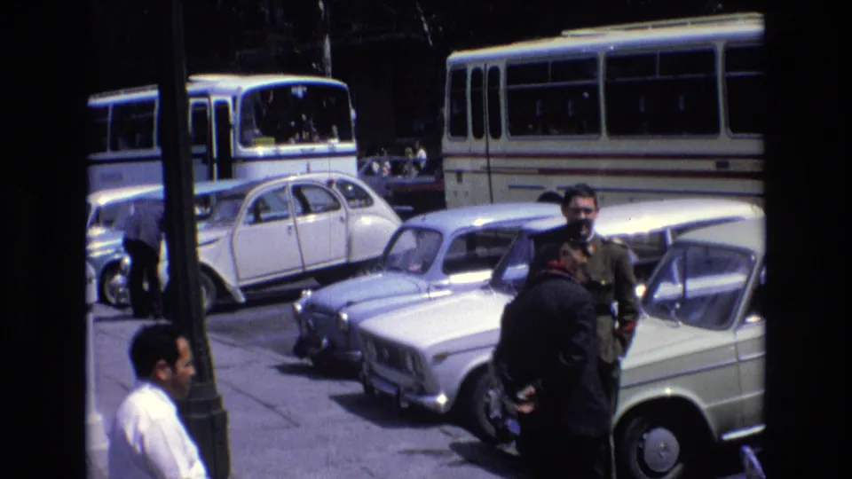1972 Spain home-movie frame showing two adult men near a row of parked classic cars, with buses and pedestrians in the background.