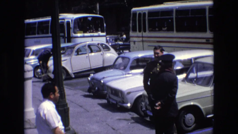 1972 Spain street frame with adult men near parked cars, including a uniformed-looking figure, with buses and a lamp post behind them.