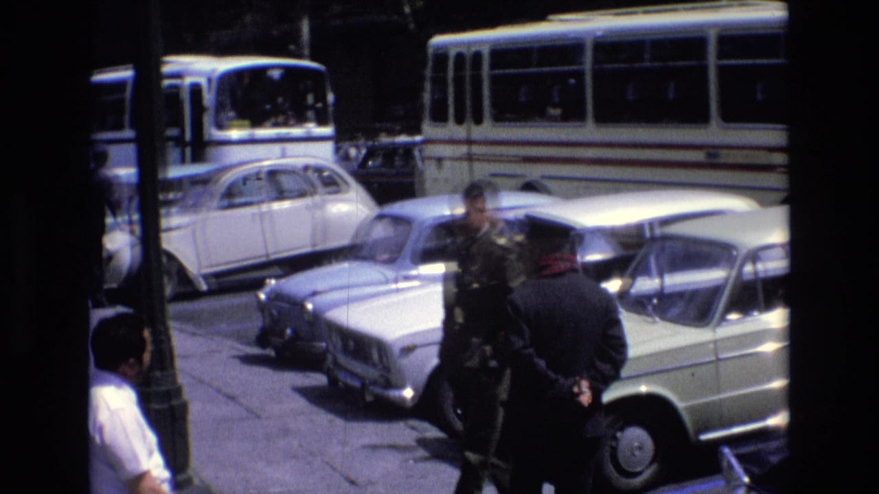 1972 Spain home-movie frame showing men near classic light-colored cars and a white public bus, partly obscured by a dark foreground edge.