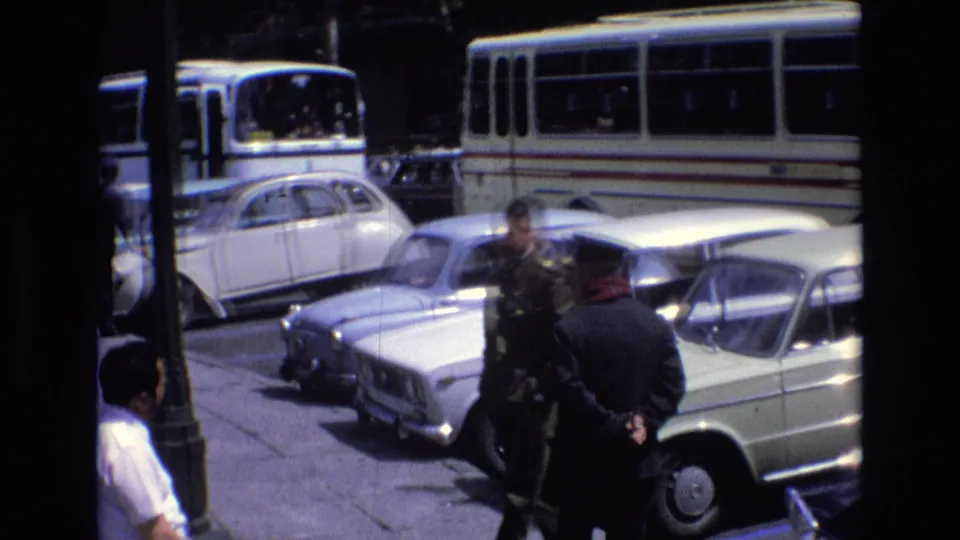 1972 Spain home-movie frame showing men near classic light-colored cars and a white public bus, partly obscured by a dark foreground edge.
