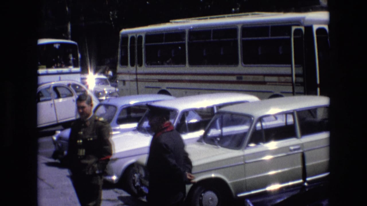 1972 Spain street view with a white bus behind parked cars and two men standing near the curb, including one in uniform-like clothing.