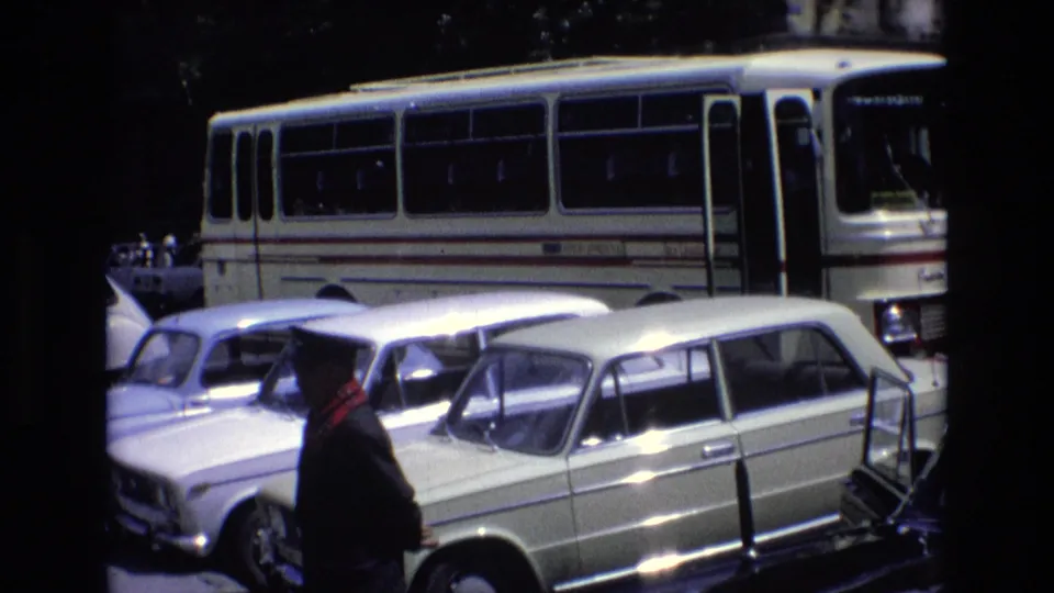 1972 Spain home-movie frame showing a man in a dark coat with a red scarf moving past light-colored parked cars in front of a white bus.