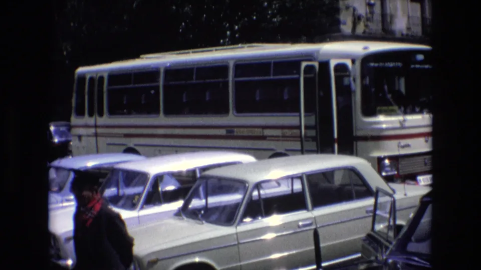 1972 Spain street-transport frame with a white public bus behind a row of classic cars while a man in dark clothing stands near the lower foreground.