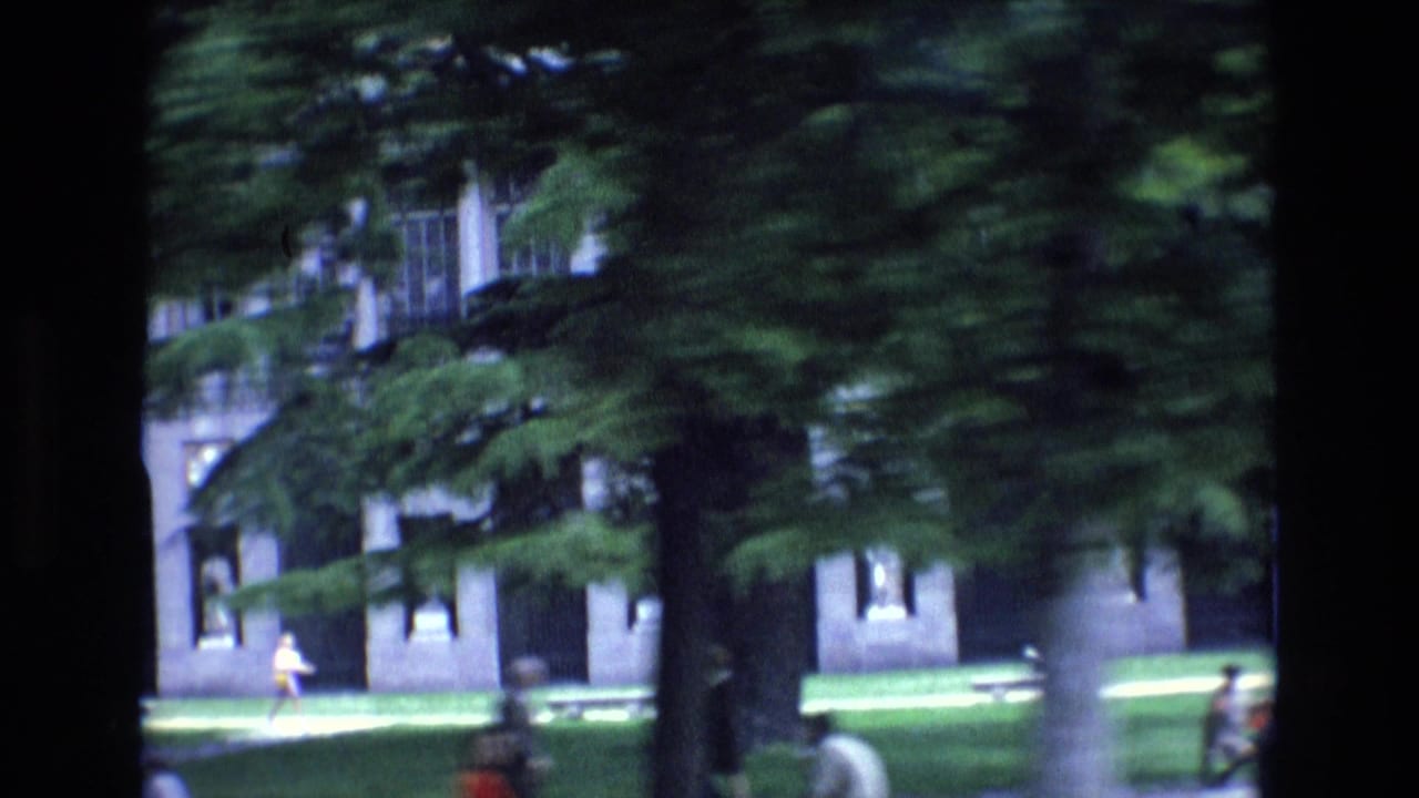 1972 Spain home-movie frame showing a leafy urban frontage with grass, benches, masonry buildings, and small pedestrians in the distance.