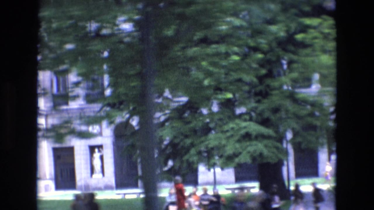 Pedestrians near trees and a pale building facade with dark doorways.