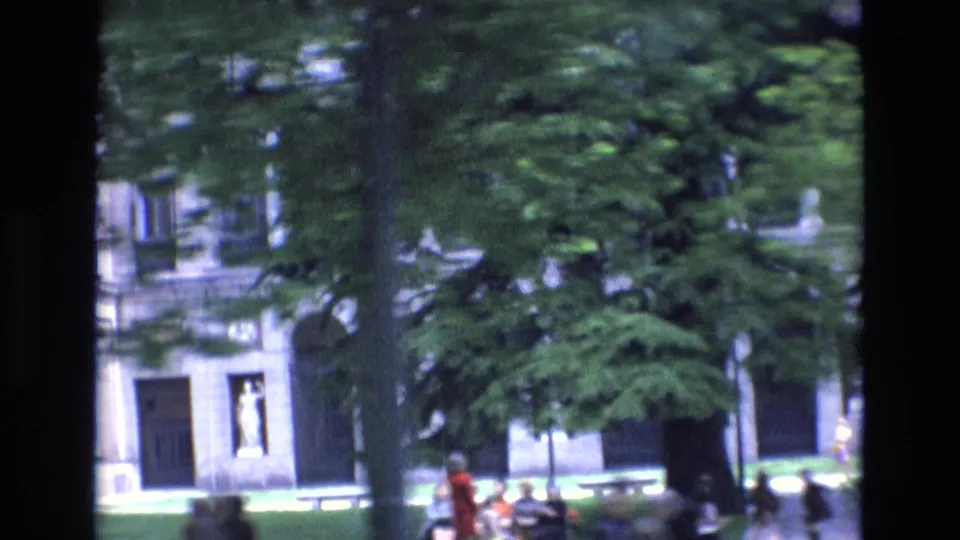 1972 Spain street-life frame with pedestrians moving near mature trees, grass, benches, and a pale urban building facade.