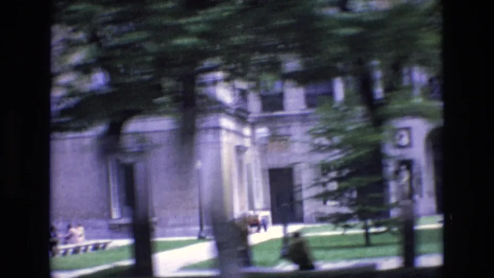 Motion-blurred 1972 Spain home-movie view of pedestrians, trees, lawn, and a light-colored building seen through foreground trunks.