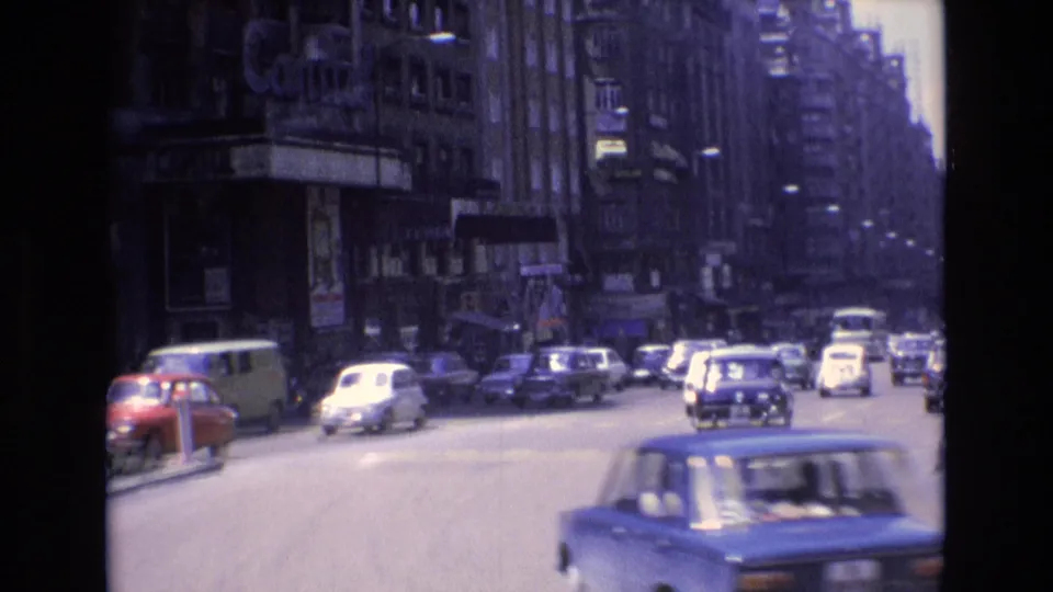 1972 Spain urban street frame showing classic cars in traffic, tall commercial buildings, storefronts, sidewalks, and pedestrians under daylight.