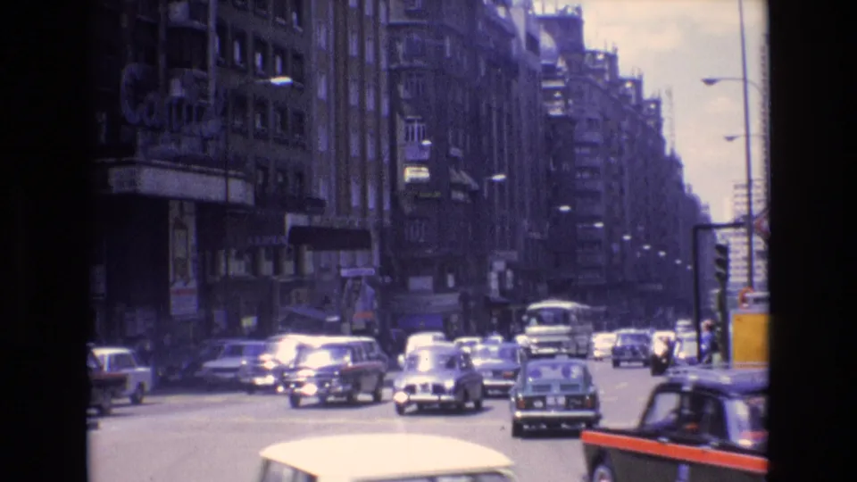 1972 Spain city traffic with a bus, compact cars, sidewalk pedestrians, and a visible Coca-Cola sign on a building along the avenue.