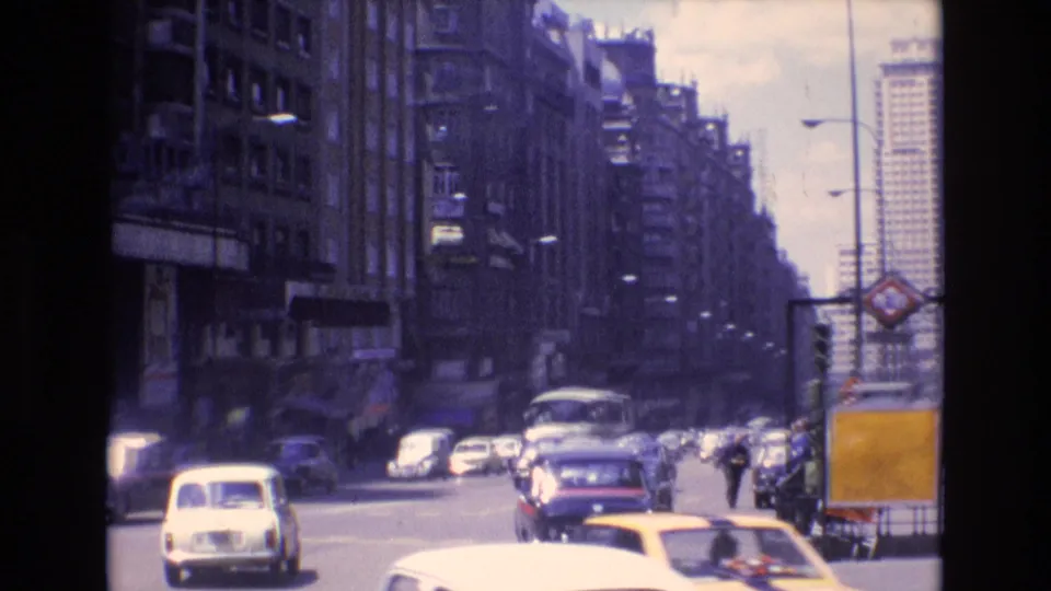 Wide 1972 Spain avenue view with classic cars, a bus, traffic signals, tall urban buildings, and a yellow curbside structure at the right edge of the frame.