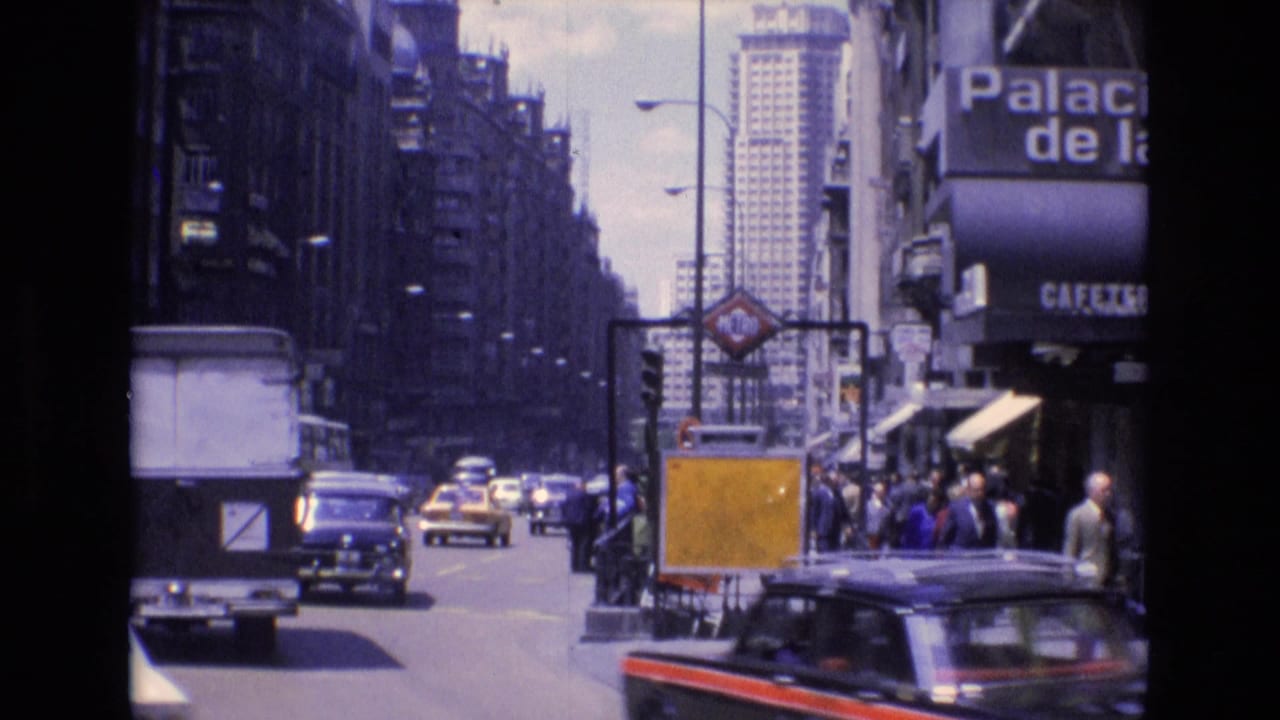 1972 Spain 8mm frame showing a busy urban street with compact cars, a white van, pedestrians along storefronts, a partial "Palacio de" sign, and a distant high-rise.