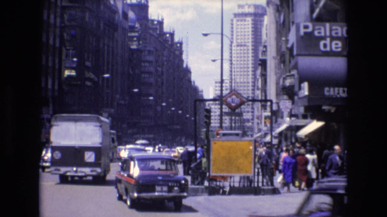 White van and dark car approach on a crowded street beside pedestrians and storefront awnings.