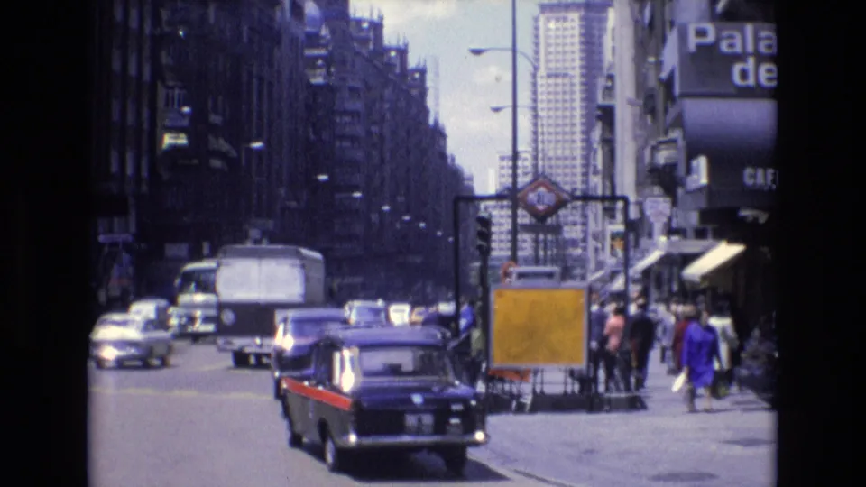 1972 Spain 8mm frame with a dark car carrying red side trim in the foreground, a white van ahead, other traffic, pedestrians near the curb, and commercial buildings lining the street.