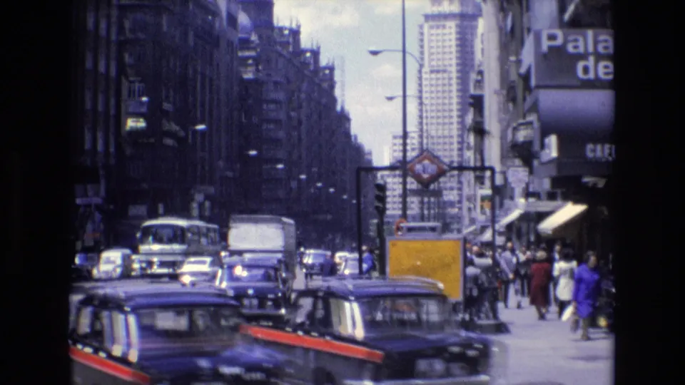 1972 Spain city street frame showing a dark foreground car with red side trim, more cars and a van in traffic, pedestrians by storefronts, a yellow panel, and a distant high-rise.