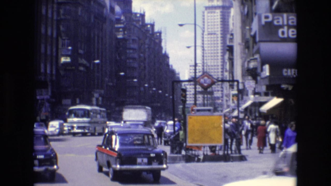 1972 Spanish city street scene with classic cars, a black taxi with red trim, buses, pedestrians, tall buildings, and a visible Metro entrance sign.