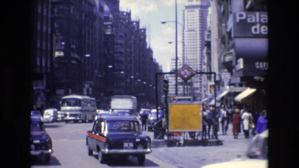 1972 Spanish city street scene with classic cars, a black taxi with red trim, buses, pedestrians, tall buildings, and a visible Metro entrance sign.