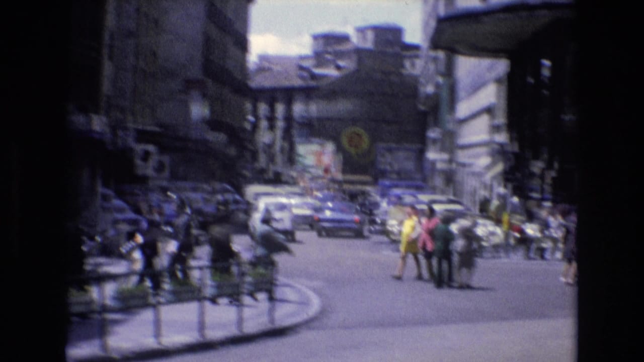 Pedestrians cross a busy intersection with cars and buildings in the background.