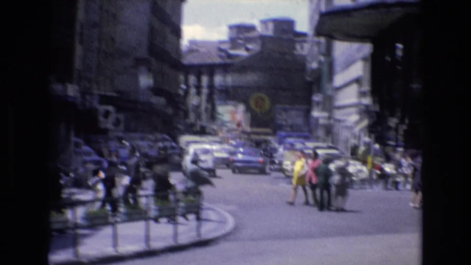 A Spanish city intersection in 1972 with pedestrians crossing, cars moving through the street, shopfronts, and dense urban buildings around the corner.