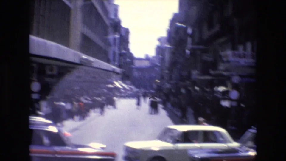 Handheld 1972 Spanish street view with motion-blurred cars, pedestrians, shop awnings, and tall buildings receding along a busy urban corridor.