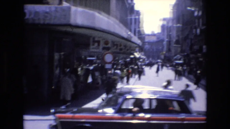 A black taxi with red trim passes in the foreground of a 1972 Spanish city street, with pedestrians, traffic signs, shop awnings, and shaded buildings behind it.
