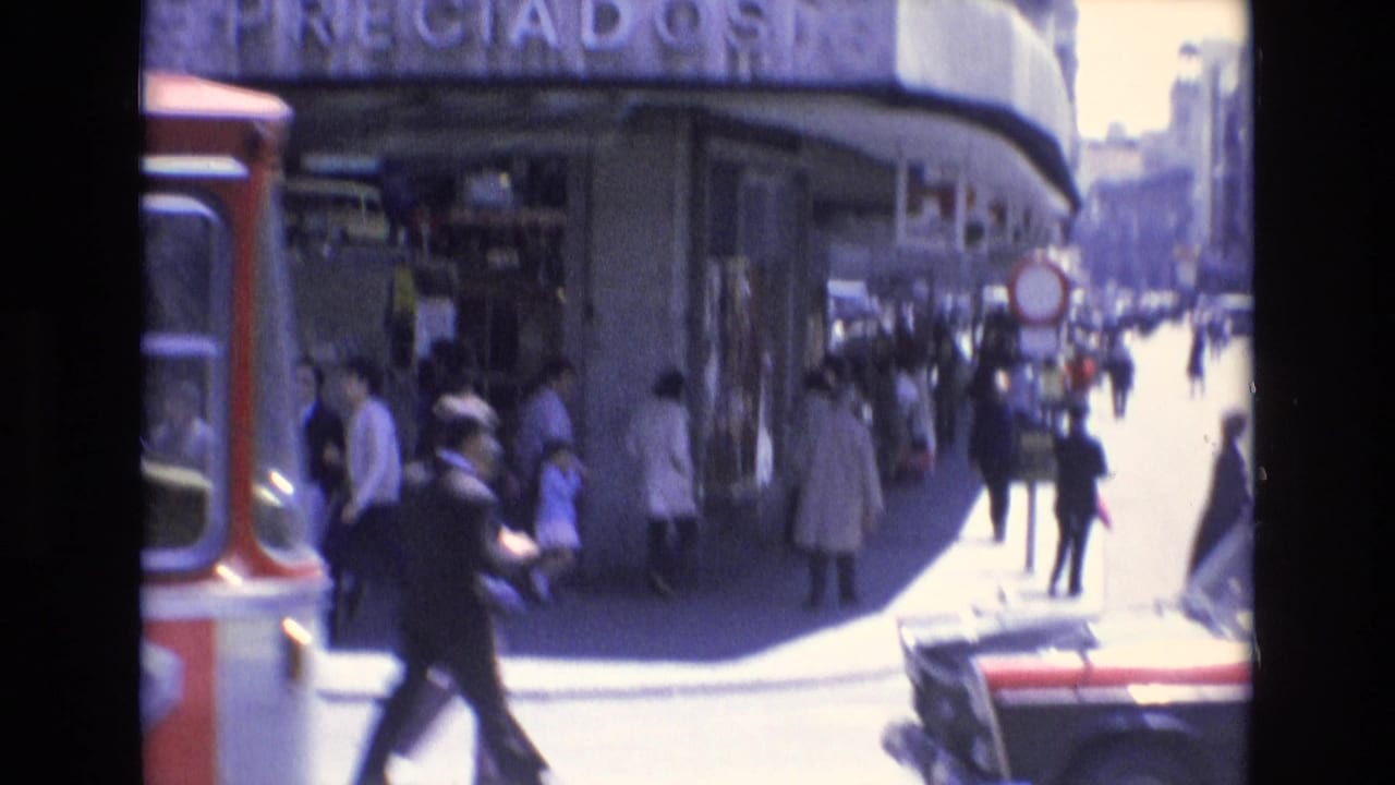 Bus, pedestrians, storefronts, and city buildings seen from a moving vehicle.