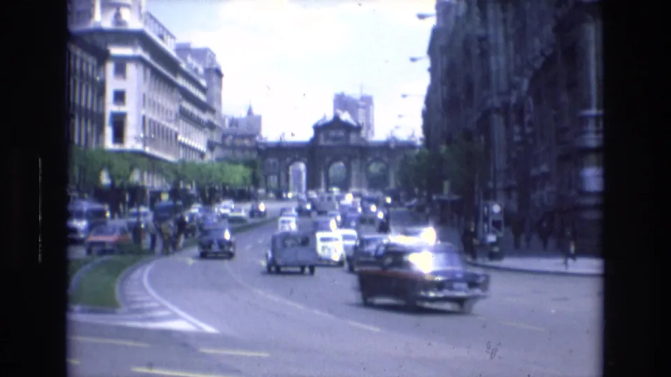 Wide 1972 Spain street view from a vehicle, with classic cars and buses moving along a curved roadway between tall buildings, trees, and a distant arched structure.