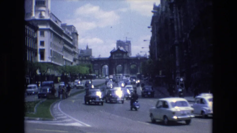 Final 8mm frame of 1972 Spain urban traffic, with compact cars, a small scooter or motorcycle, broad roadway markings, tall buildings, trees, and a distant arched structure.