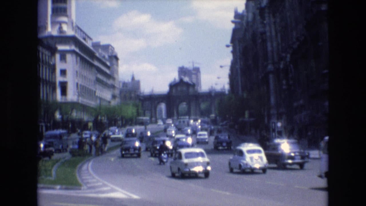 1972 Spain 8mm frame showing cars and small vehicles moving along a curved urban roadway toward a distant arched gateway between multi-story buildings.