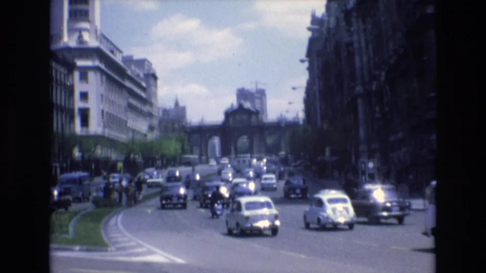 1972 Spain 8mm frame showing cars and small vehicles moving along a curved urban roadway toward a distant arched gateway between multi-story buildings.