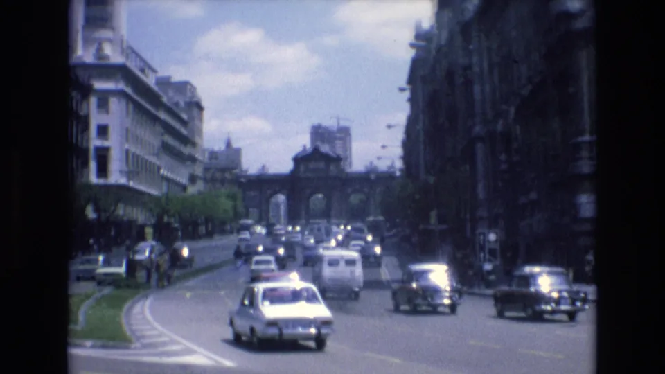 1972 Spain 8mm frame with a compact white car in the foreground, other vehicles beyond it, and a distant arched gateway at the end of the urban street.
