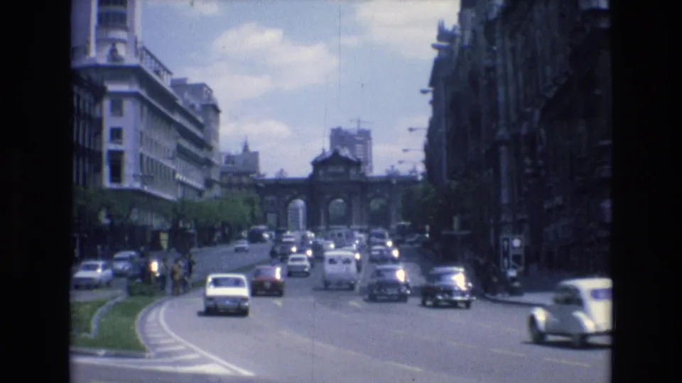 1972 Spain home-movie frame showing several cars on a broad urban roadway, pedestrians near the street edge, and a distant arched gateway framed by dense city buildings.