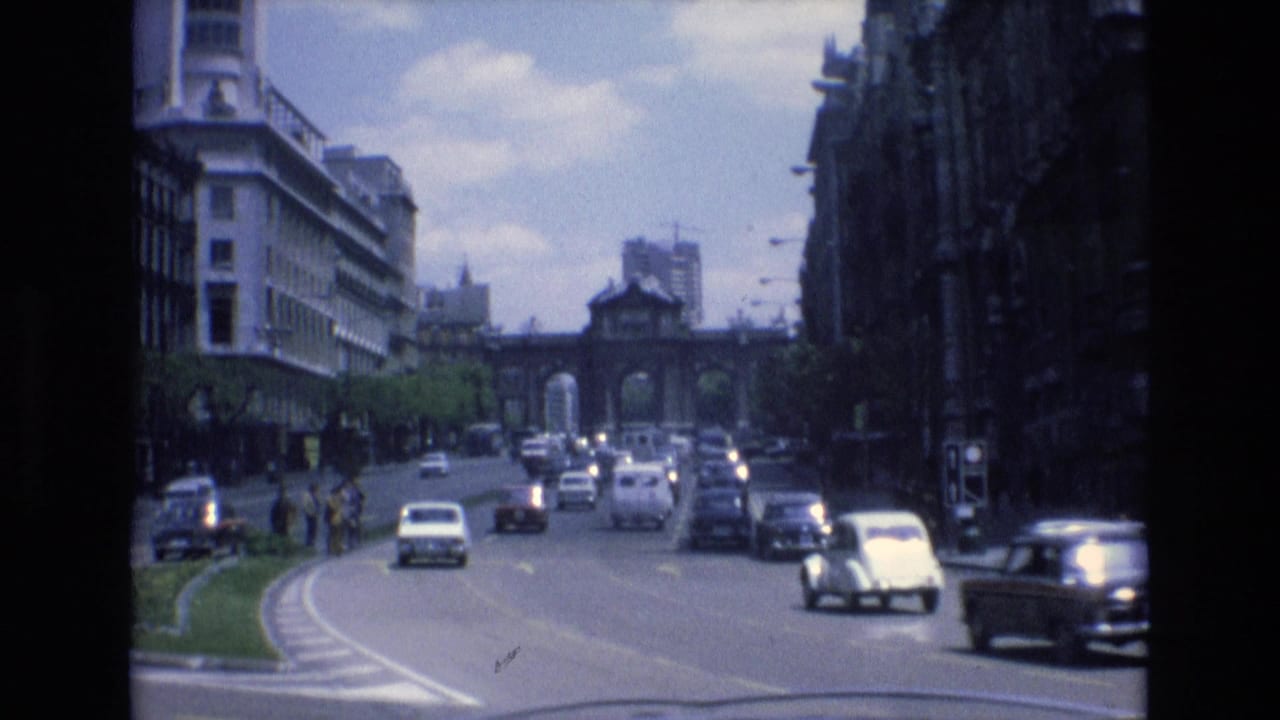 1972 Spain 8mm street view with classic cars traveling along a curved urban roadway, pedestrians near the sidewalk, tall buildings, and a distant arched gateway.