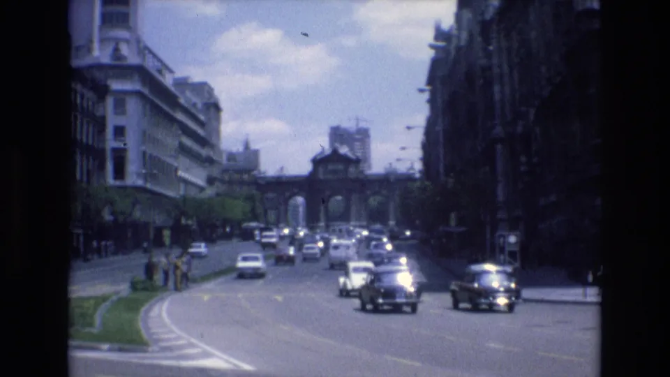 1972 Spain street traffic with several mid-century cars moving through a broad urban avenue, pedestrians at the left edge, and historic architecture visible beyond the road.